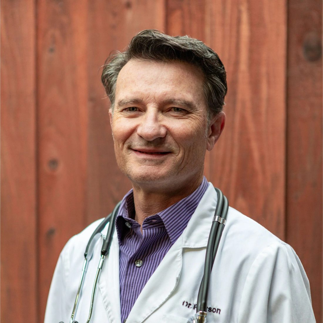Smiling male veterinarian in white coat with stethoscope against wood wall